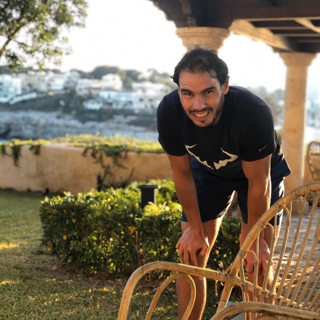 "رافاييل نادال يبتسم أثناء وقوفه في حديقة بمنزله في مايوركا، مرتديًا قميصًا رياضيًا غير رسمي في لحظة استرخاء."